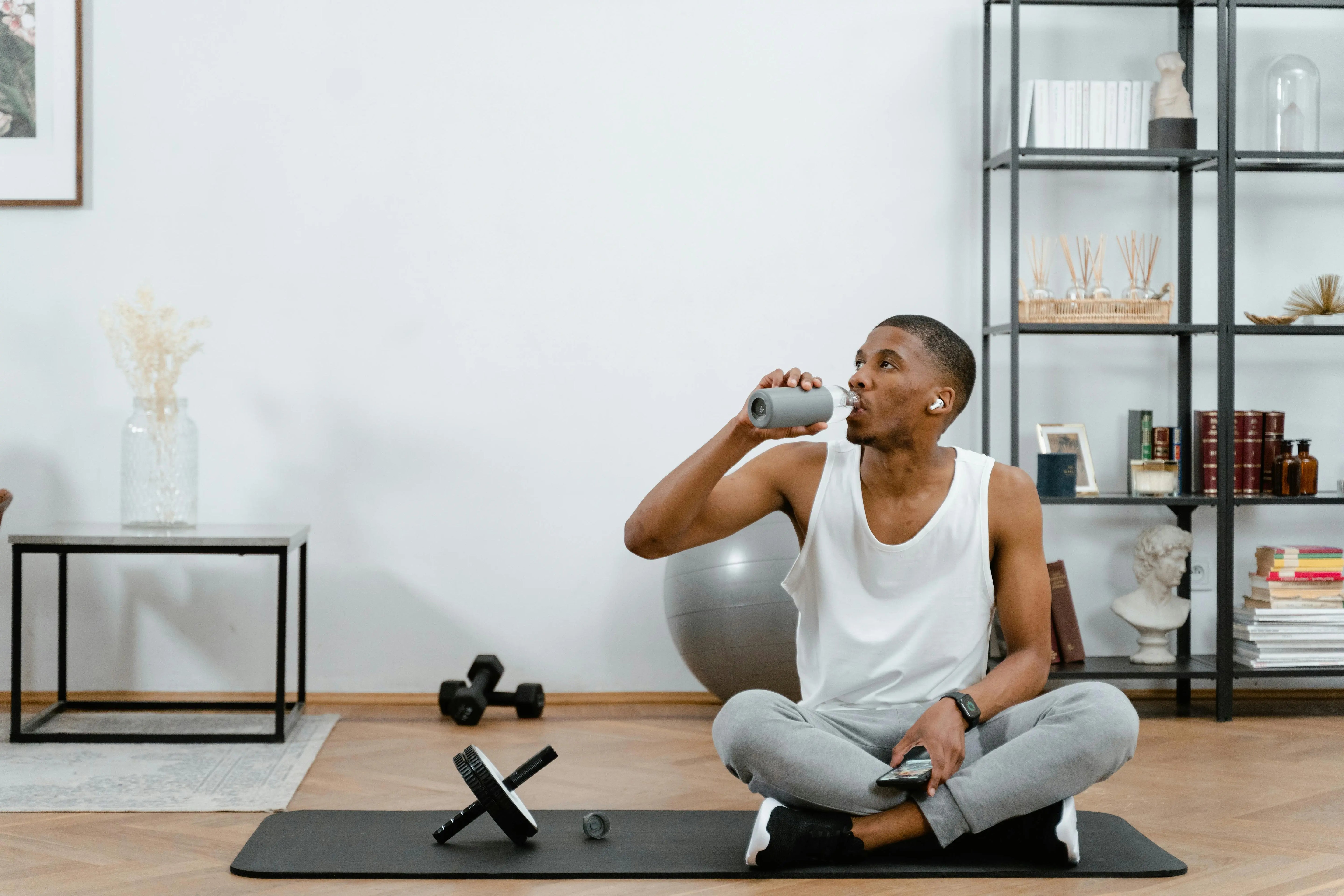 Person sitting on a yoga mat in a home setting, drinking from a bottle.